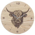 A round wooden wall clock with a natural light wood grain finish, featuring a dark engraved illustration of a Highland cow at the center and dark brown Arabic numerals marking the hours.