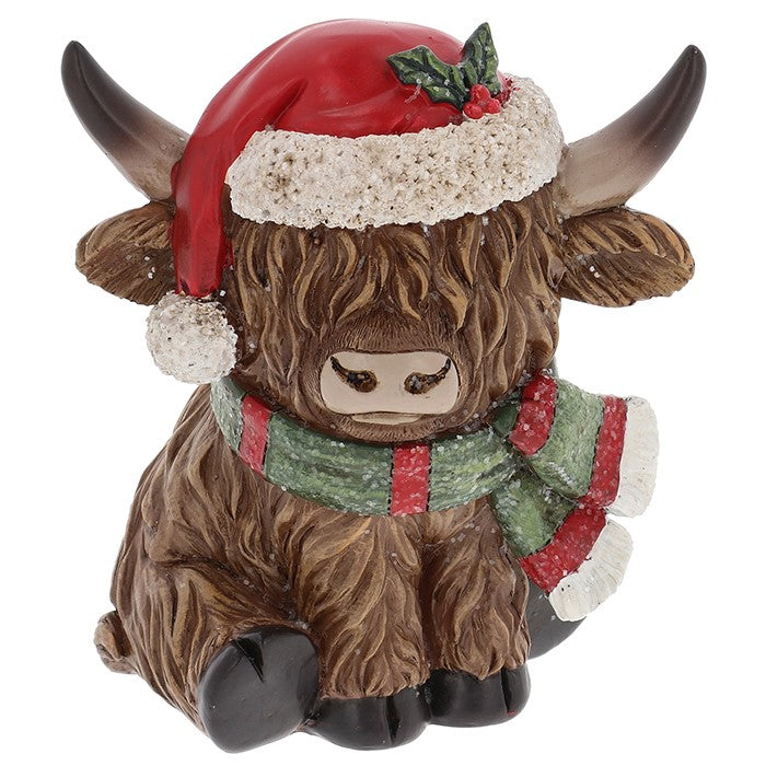 A Highland cow figurine wearing a red Santa hat with a white fluffy trim and holly sprig, along with a green and red striped scarf, set against a plain white background.
